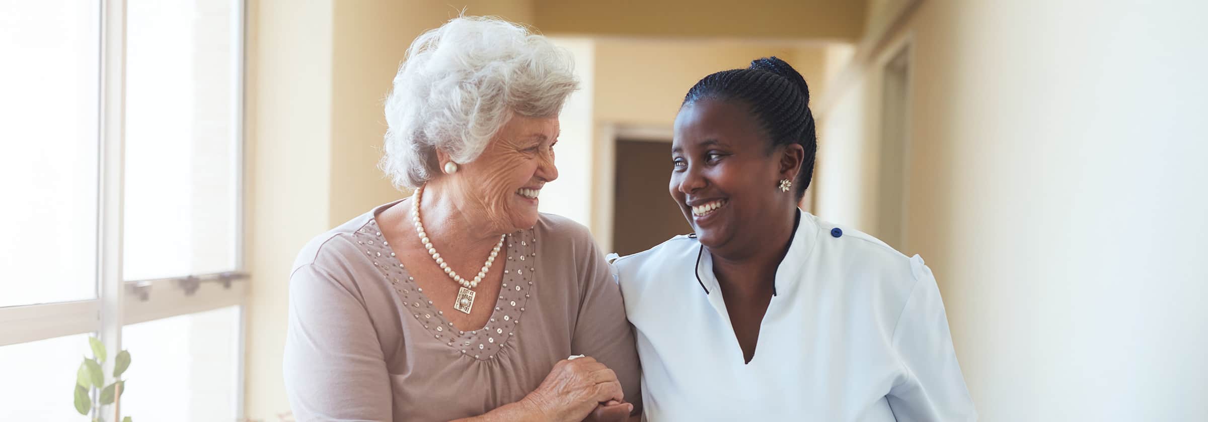 A female caregiver holds an older woman's arm while walking down a hall.