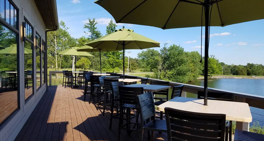 Wide shot of Traditions of Columbus back deck with outdoor furniture of tables, green umbrellas, and wooden chairs overlooking the lake.