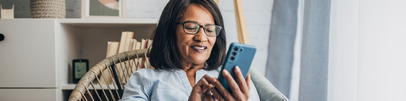 A senior woman sitting in a wooden chair smiling and looking down while scrolling on her blue smartphone