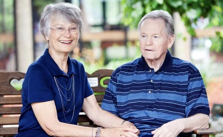Elderly couple sitting outside together holding hands, smiling for the camera, and enjoying nature together. At Traditions of Columbus in Columbus, Indiana.