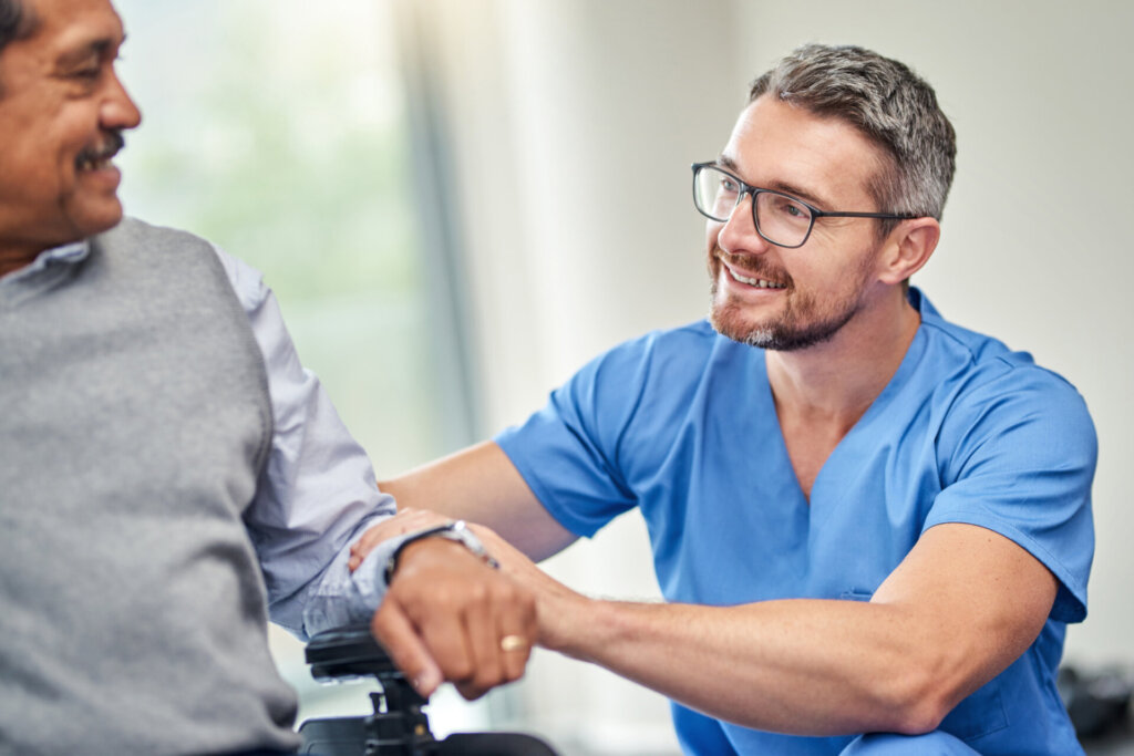 A male caregiver, wearing blue scrubs, holds onto an elderly man's arm as he sits in a wheelchair. They are having a pleasant conversation and enjoying the afternoon. At Traditions of Columbus in Columbus, Indiana.